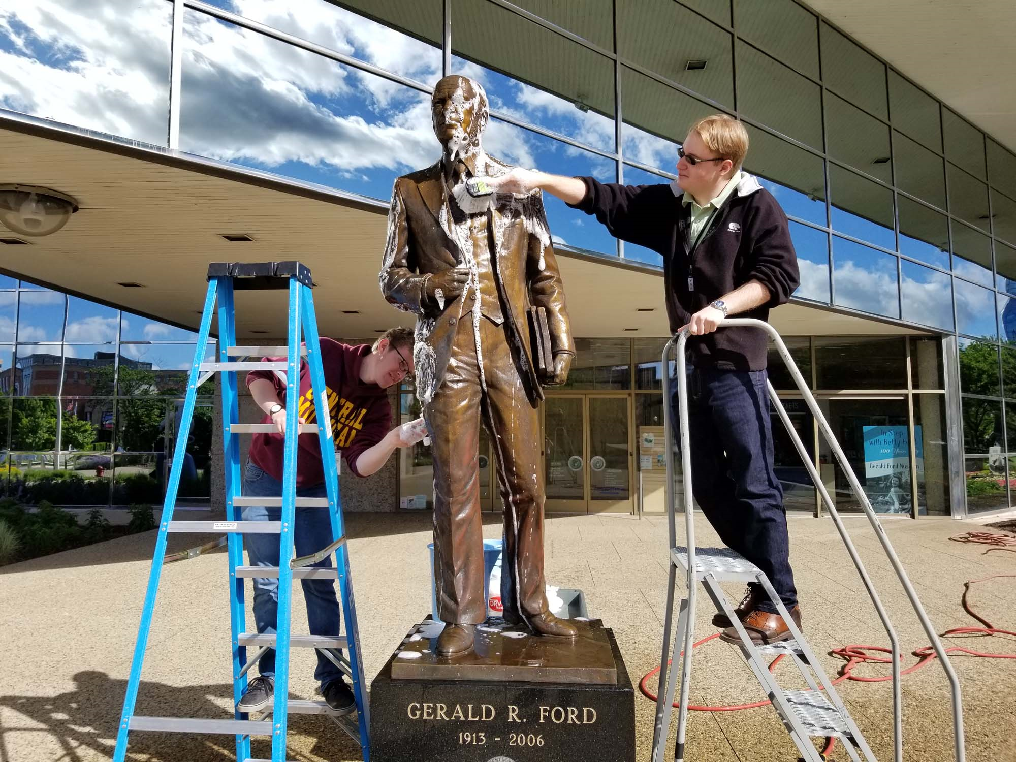 A man cleans a statue of a man covered in soap with a brush while on a ladder.  